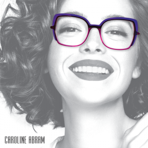 Black-and-white photo of a smiling woman wearing bold square purple-and-pink glasses, Caroline Abram